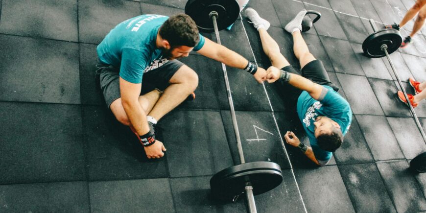 two men relaxing on the gym floor tired after strength training fist bump each other